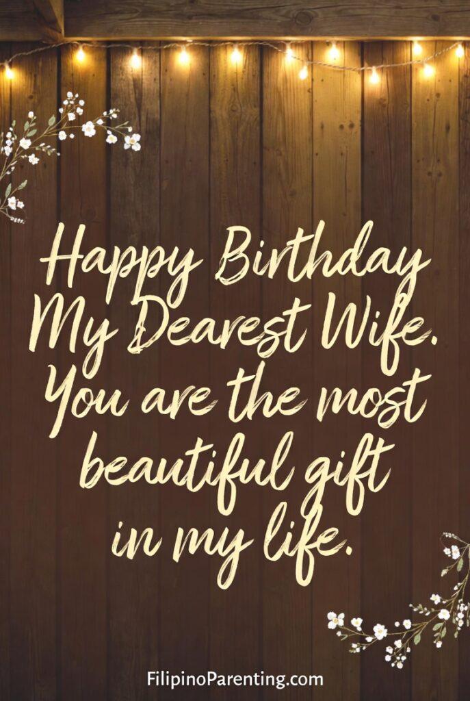 Maligayang Kaarawan Asawang Babae: 25 Heartfelt Birthday Messages, Prayers, and Quotes Celebrate your spouse with this cozy and rustic 'Happy Birthday My Dearest Wife' digital card. Set against a warm wooden background with glowing string lights and elegant white flowers, the message reads: 'You are the most beautiful gift in my life.' This heartfelt design is perfect for husbands looking to share a sincere, romantic birthday tribute on social media or as a personalized message to honor their wife’s special day.