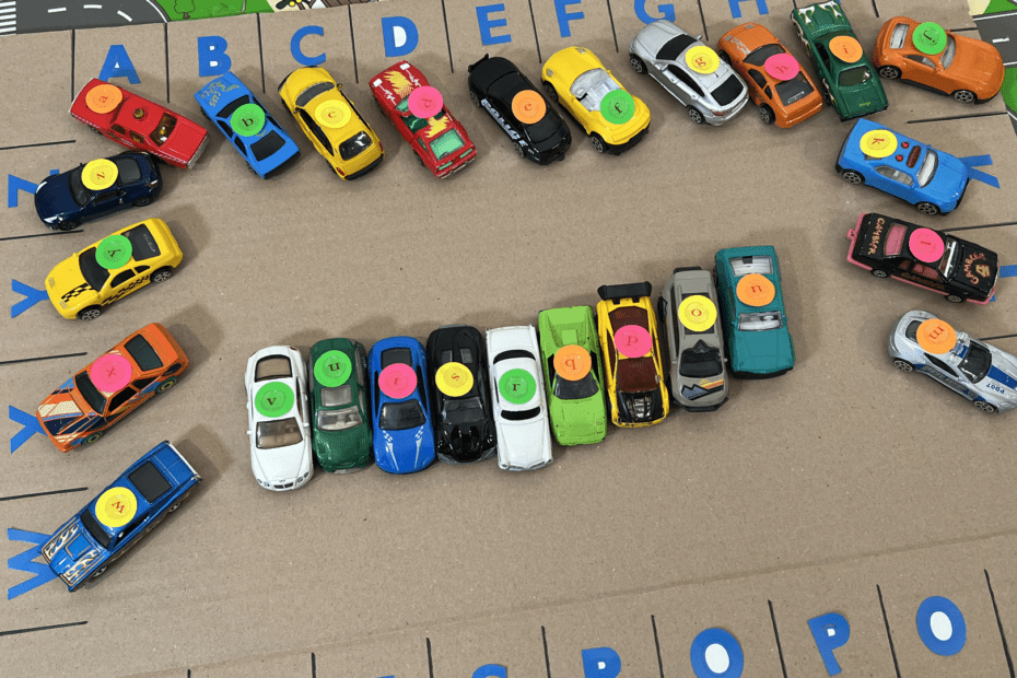 Angled view of toy vehicles with lowercase alphabet stickers parked on a handmade cardboard letter matching mat