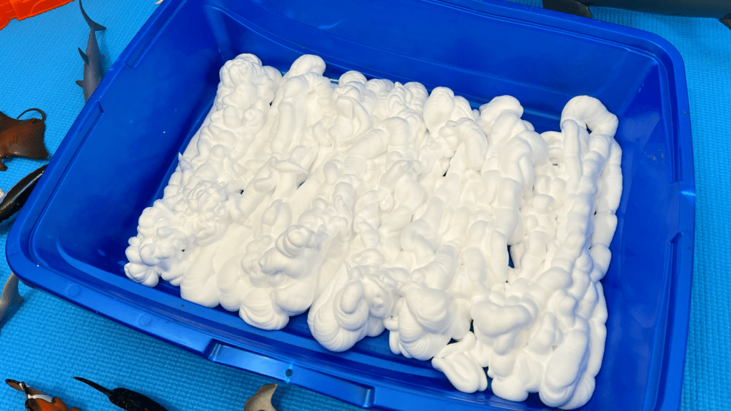 Easy Shaving Cream Sea Foam Sensory Bin for Home Learning A blue plastic tub filled with rows of fluffy white shaving cream foam, set on a blue play mat with toy sea creatures surrounding it.