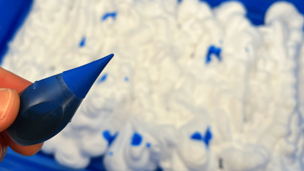 Easy Shaving Cream Sea Foam Sensory Bin for Home Learning A close-up of a hand holding a blue food coloring bottle over a bed of white shaving cream in a sensory bin.