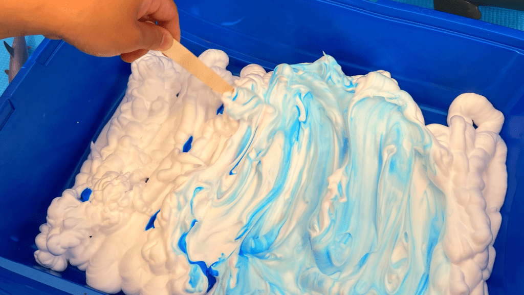 Easy Shaving Cream Sea Foam Sensory Bin for Home Learning A hand using a wooden craft stick to swirl blue coloring into fluffy white shaving cream inside a blue plastic sensory bin.