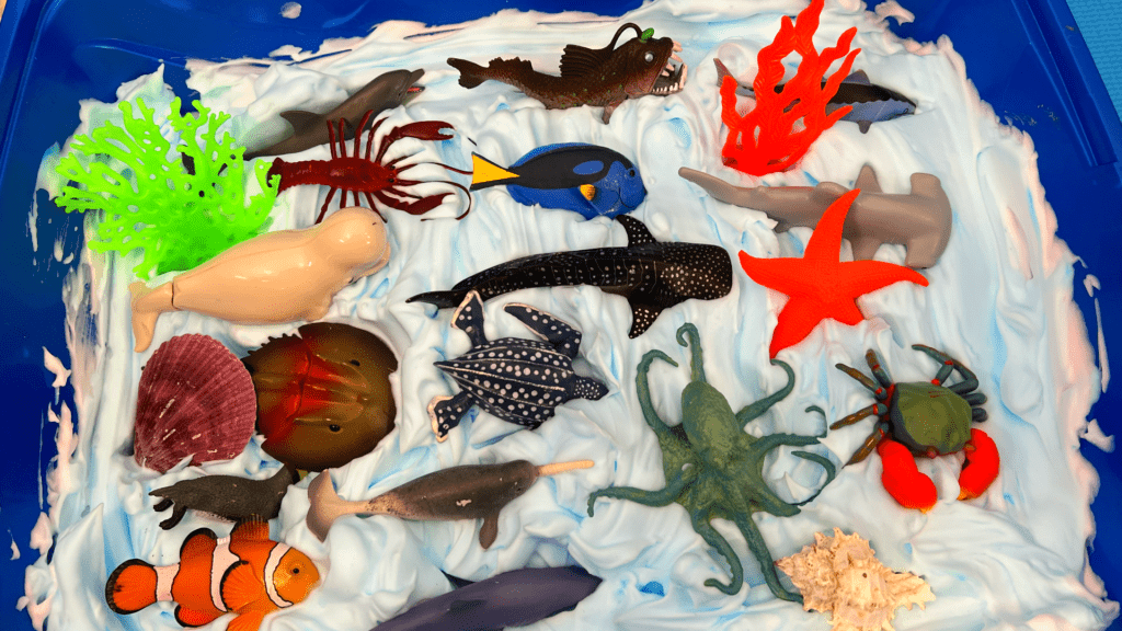 Easy Shaving Cream Sea Foam Sensory Bin for Home Learning A high-angle view of various toy ocean creatures, including an octopus, sea turtle, crab, and starfish, arranged on top of blue-swirled shaving cream foam inside a sensory tub.
