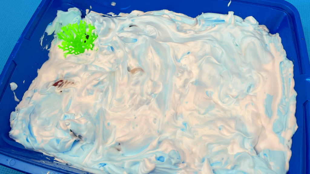 Easy Shaving Cream Sea Foam Sensory Bin for Home Learning A blue plastic sensory bin filled with swirled white and light-blue shaving cream foam and a bright green plastic coral piece.