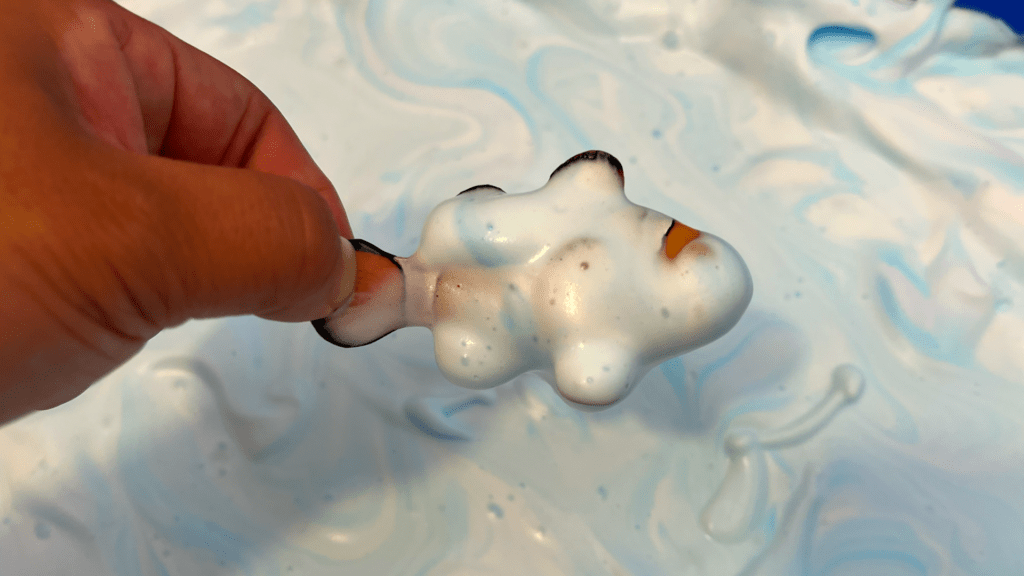 Easy Shaving Cream Sea Foam Sensory Bin for Home Learning A hand holding a toy clownfish covered in blue-swirled shaving cream foam above an ocean-themed sensory bin.