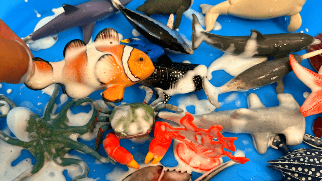Easy Shaving Cream Sea Foam Sensory Bin for Home Learning A hand placing a toy clownfish into a blue water rinse tub alongside other toy sea creatures covered in shaving cream foam.